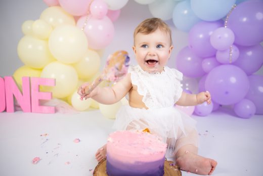 Baby playfully smashing cake with a wooden spoon and rainbow balloons.