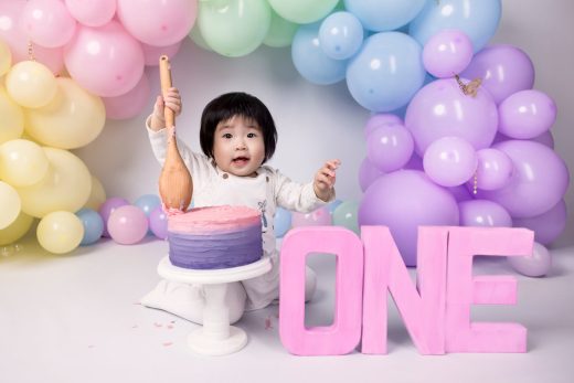 Baby playfully smashing cake with a wooden spoon and rainbow balloons.