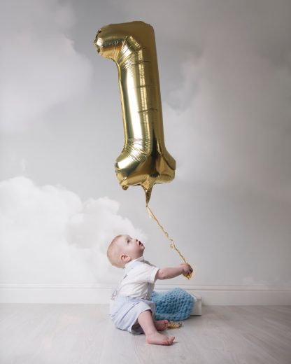 A young boy looks up at gold one balloon with a cloudy background.