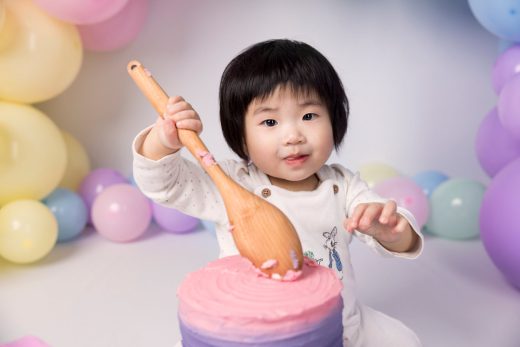 Baby playfully smashing cake with a wooden spoon and rainbow balloons.