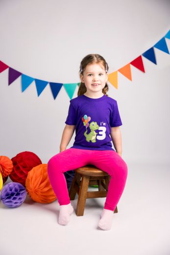 Three year old girl sits on stool with bunting and rainbow decorations in the background.