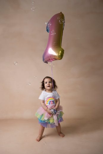 One year old girl in a rainbow tutu standing with rainbow balloon on a beige background.